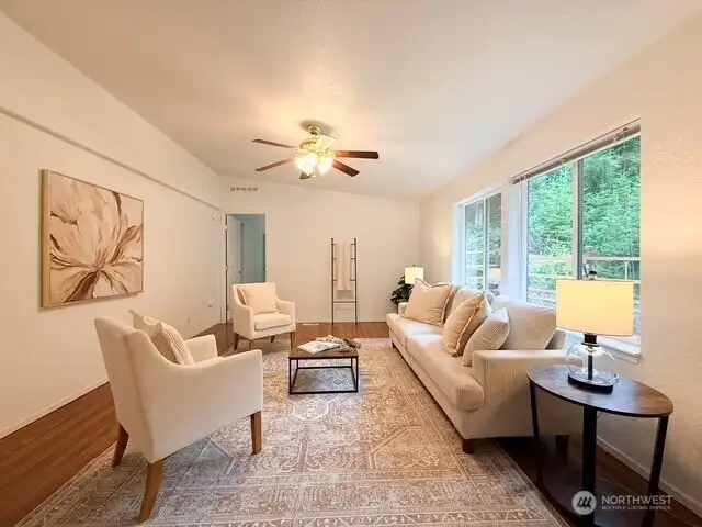 This bright and welcoming living room features vaulted ceilings, a cooling ceiling fan, warm wood-look flooring, and massive windows that flood the space with natural light while framing the serene forest views.