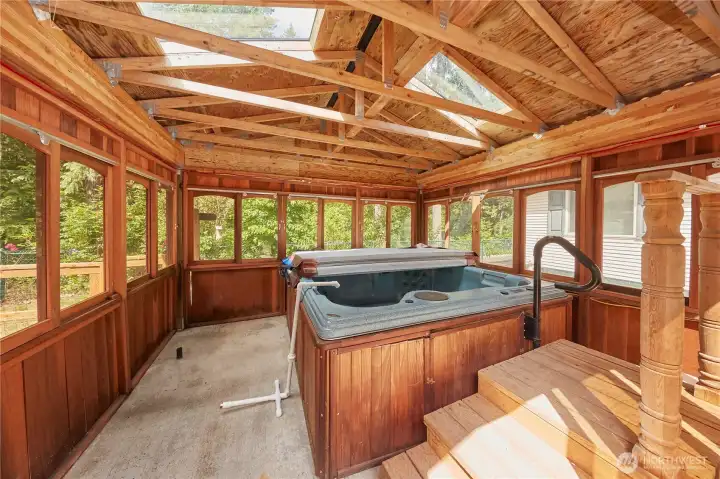 Relax year-round in the private, enclosed hot tub room, featuring rustic wood detailing, vaulted ceilings with skylights, and expansive wrap-around windows looking out to the forest.