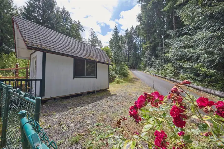A charming exterior view of the detached cottage-style outbuilding, nestled in a forest setting and framed by beautifully blooming red roses.