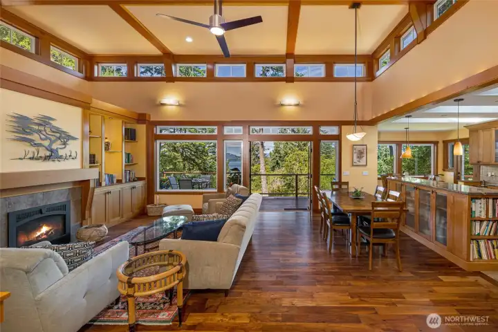 Soaring coffered ceilings with rich wood beams frame this luminous great room, where a wall of windows and French doors open to a deck overlooking the treetops and sparkling San Juan Island waters. The seamless open-plan layout connects the living area—anchored by a stone fireplace and built-in shelving—to the formal dining and kitchen beyond. Soaring coffered ceilings with rich wood beams frame this luminous great room, where a wall of windows and French doors open to a deck overlooking the treetops and sparkling San Juan Island waters. The seamless open-plan layout connects the living area—anchored by a stone fireplace and built-in shelving—to the formal dining and kitchen beyond.