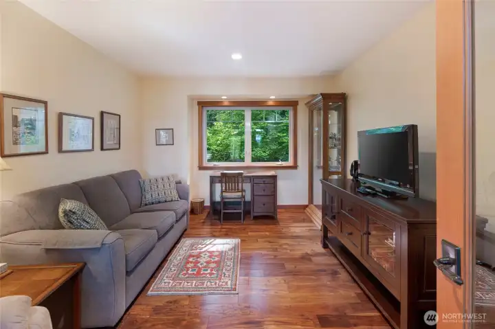 Warm and versatile den features rich cherry hardwood flooring, a sleeper sofa, and a substantial dark wood media console with flat-screen TV, making it ideal as a home office, guest suite, or entertainment room. Custom wood-trimmed windows complete this refined, multi-purpose space. Warm and versatile den features rich cherry hardwood flooring, a sleeper sofa, and a substantial dark wood media console with flat-screen TV, making it ideal as a home office, guest suite, or entertainment room. Custom wood-trimmed windows complete this refined, multi-purpose space.