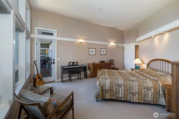 The generously proportioned primary bedroom offers a relaxed sitting area to enjoy water views and direct access to an outdoor deck through a glass-paned door, creating seamless indoor-outdoor living. High ceilings, warm neutral tones and thoughtfully placed lighting lend an airy, resort-like ambiance throughout. The generously proportioned primary bedroom offers a relaxed sitting area to enjoy water views and direct access to an outdoor deck through a glass-paned door, creating seamless indoor-outdoor living. High ceilings, warm neutral tones and thoughtfully placed lighting lend an airy, resort-like ambiance throughout.
