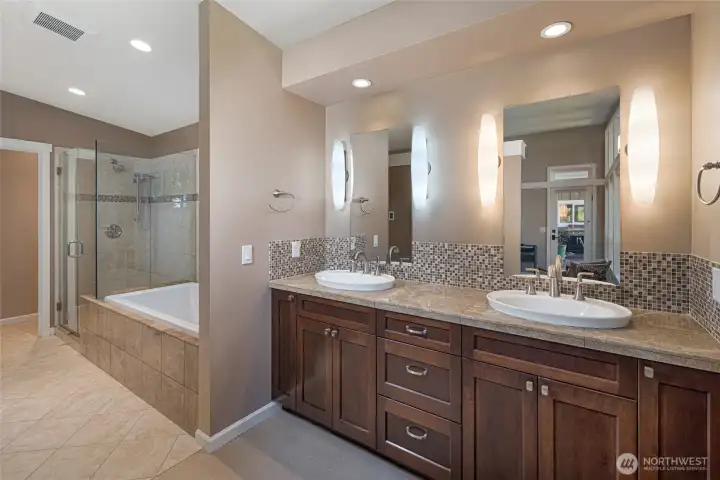 The luxurious primary en-suite bath features a long dual-sink vanity with vessel bowls, granite countertops, rich dark cabinetry, and a stunning mosaic tile backsplash accented by elegant oval sconce lighting. The spa-inspired design is completed by a glass-enclosed walk-in shower and a deep soaking tub just steps away. The luxurious primary en-suite bath features a long dual-sink vanity with vessel bowls, granite countertops, rich dark cabinetry, and a stunning mosaic tile backsplash accented by elegant oval sconce lighting. The spa-inspired design is completed by a glass-enclosed walk-in shower and a deep soaking tub just steps away.