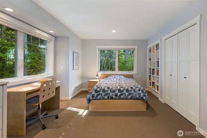 A bright and well-proportioned secondary bedroom features large windows with tranquil forest views, a built-in desk area perfect for a student or remote worker, and a handsome white built-in bookcase flanking a generous closet. Recessed lighting and neutral tones make this a versatile and welcoming space. A bright and well-proportioned secondary bedroom features large windows with tranquil forest views, a built-in desk area perfect for a student or remote worker, and a handsome white built-in bookcase flanking a generous closet. Recessed lighting and neutral tones make this a versatile and welcoming space.