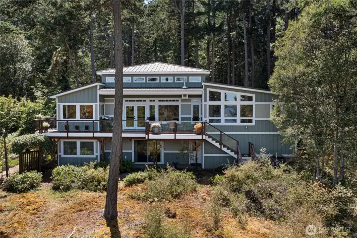 The dramatic view-facing rear elevation showcases a broad upper-level deck with glass-panel and wood railings, expansive clerestory windows, and a lower guest accommodations—all oriented to capture sweeping water and island vistas from both levels. The dramatic view-facing rear elevation showcases a broad upper-level deck with glass-panel and wood railings, expansive clerestory windows, and a lower guest accommodations—all oriented to capture sweeping water and island vistas from both levels.