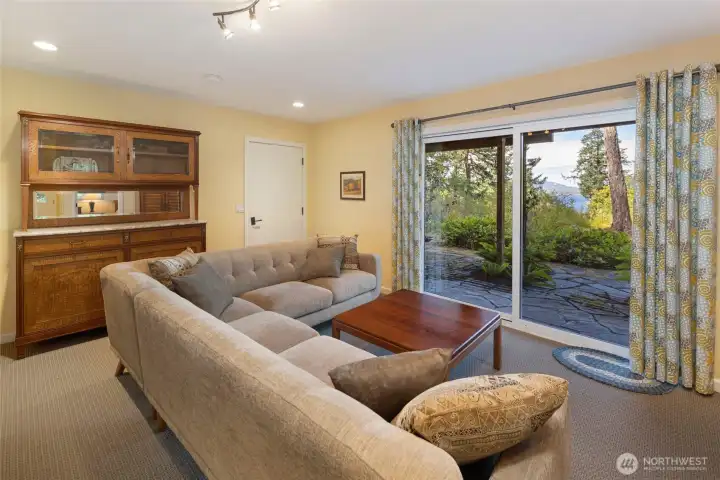 Bright and welcoming first floor guest living area features a large sliding glass door that opens to a lush stone patio and water views, seamlessly blending indoor comfort with the verdant Pacific Northwest landscape. Track lighting and recessed fixtures illuminate the space, with a a glass paneled door inviting you to an enchanting deck and ornamental garden beyond. Bright and welcoming first floor guest living area features a large sliding glass door that opens to a lush stone patio and water views, seamlessly blending indoor comfort with the verdant Pacific Northwest landscape. Track lighting and recessed fixtures illuminate the space, with a a glass paneled door inviting you to an enchanting deck and ornamental garden beyond.