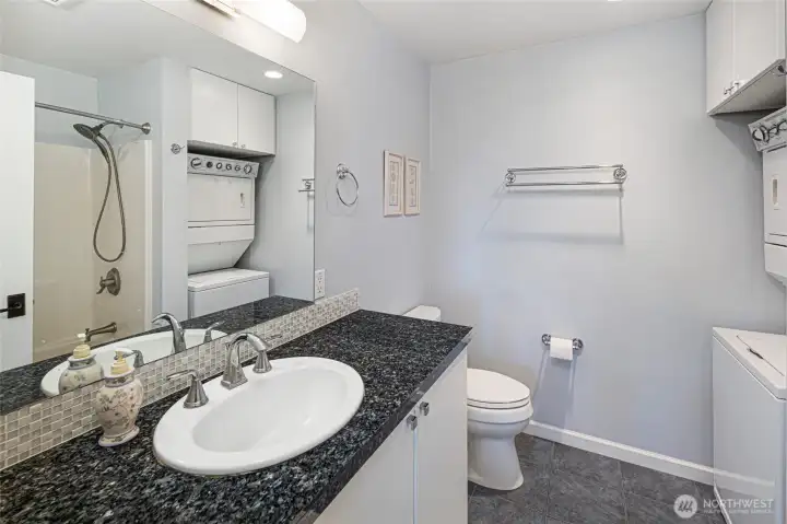 Well-appointed full bathroom in the guest quarters features blue pearl granite countertops, a tub/shower, and a stacked washer-dryer conveniently integrated into the same space for effortless single-level living. Clean white cabinetry, chrome fixtures, and mosaic tile accents lend a polished, cohesive aesthetic. Well-appointed full bathroom in the guest quarters features blue pearl granite countertops, a tub/shower, and a stacked washer-dryer conveniently integrated into the same space for effortless single-level living. Clean white cabinetry, chrome fixtures, and mosaic tile accents lend a polished, cohesive aesthetic.