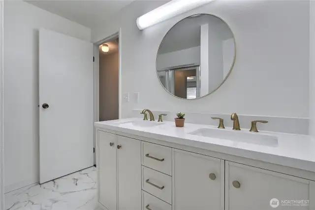 Stylish and updated, this guest bathroom features a sleek double vanity with quartz countertops, brushed gold fixtures, and a modern round mirror. A fresh white palette and durable vinyl flooring give the space a clean and contemporary look, perfect for everyday use or guests.