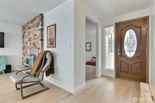 Step inside to a light and welcoming entry with beautiful new wide-plank hardwood laminate flooring and a decorative glass-inset front door that adds charm and character. Natural light pours in from the adjacent sidelight window, highlighting the fresh interior paint and clean, modern lines. A cozy peek into the front sitting room and a glimpse of the brick-accented fireplace create a feeling of home right from the start.