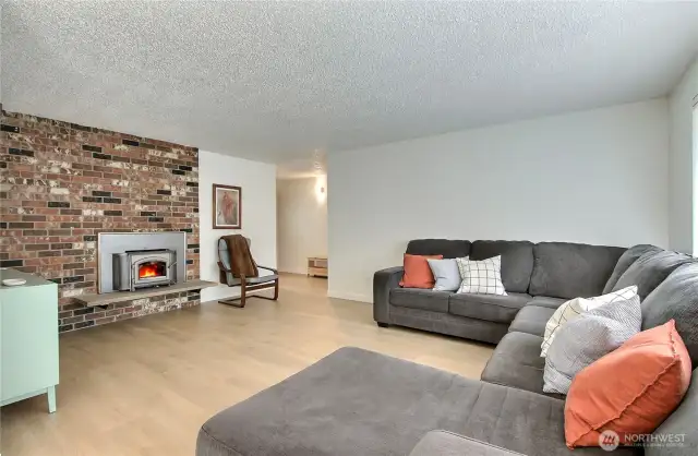 Spacious and light-filled living room features stunning wide-plank laminate flooring and a cozy wood-burning fireplace with a striking full brick surround. The open layout offers plenty of room for entertaining or relaxing, with large windows bringing in natural light. A warm and welcoming centerpiece to the home!