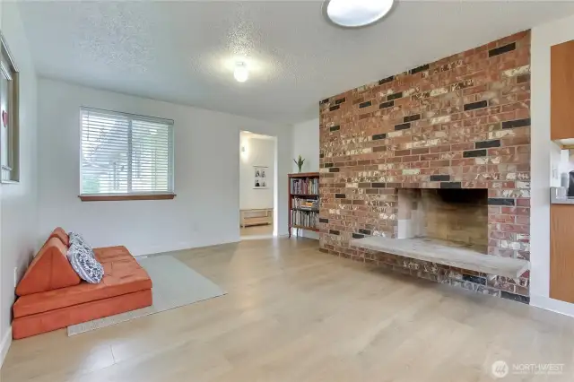 Inviting family room featuring brand new wide-plank flooring and a bold brick fireplace that anchors the space with warmth and character. The front-facing window adds a pleasant street view, while the open layout offers endless possibilities for everyday living or entertaining.