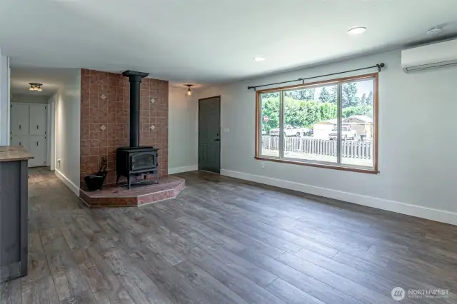 Family room looking back to front door showing wood stove and mini-split system. Natural lighting with big windows.