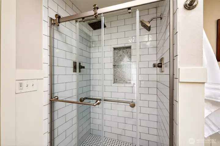 The updated primary shower is finished to a hotel standard — floor-to-ceiling subway tile, a herringbone mosaic niche, dual showerheads including an overhead rain fixture, and frameless sliding glass doors.