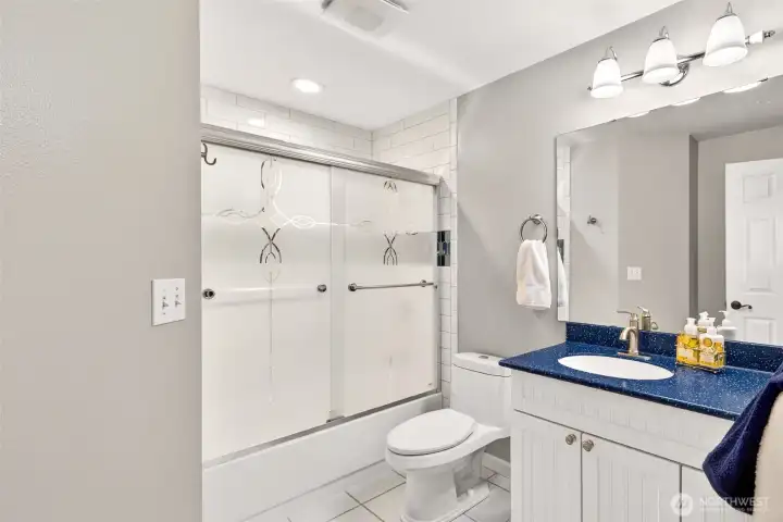 The full hall bath serves the secondary bedrooms with a clean, functional layout — white cabinetry, a bold blue seamless countertop, subway tile tub surround, and decorative sliding glass doors add personality and polish.