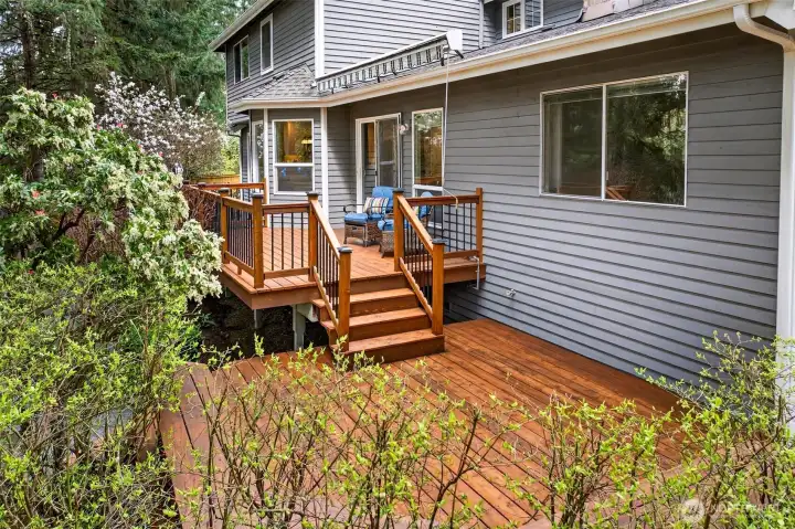 The multi-level deck with staircase connection showcases the home's thoughtful outdoor design — seamlessly linking the main living level to the yard and patio below, all framed by blooming ornamentals and mature evergreens.