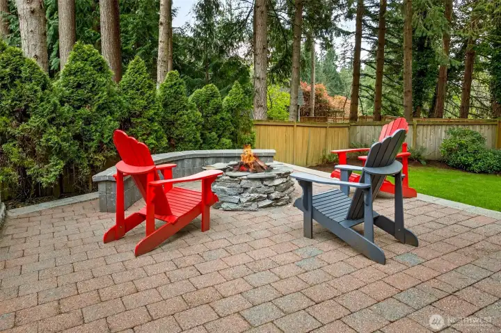 The paver patio fire pit area is an entertainer's dream — a beautifully designed outdoor living room with a built-in stone surround fire pit, curved seat wall, and a private, fully fenced backdrop of towering evergreens.