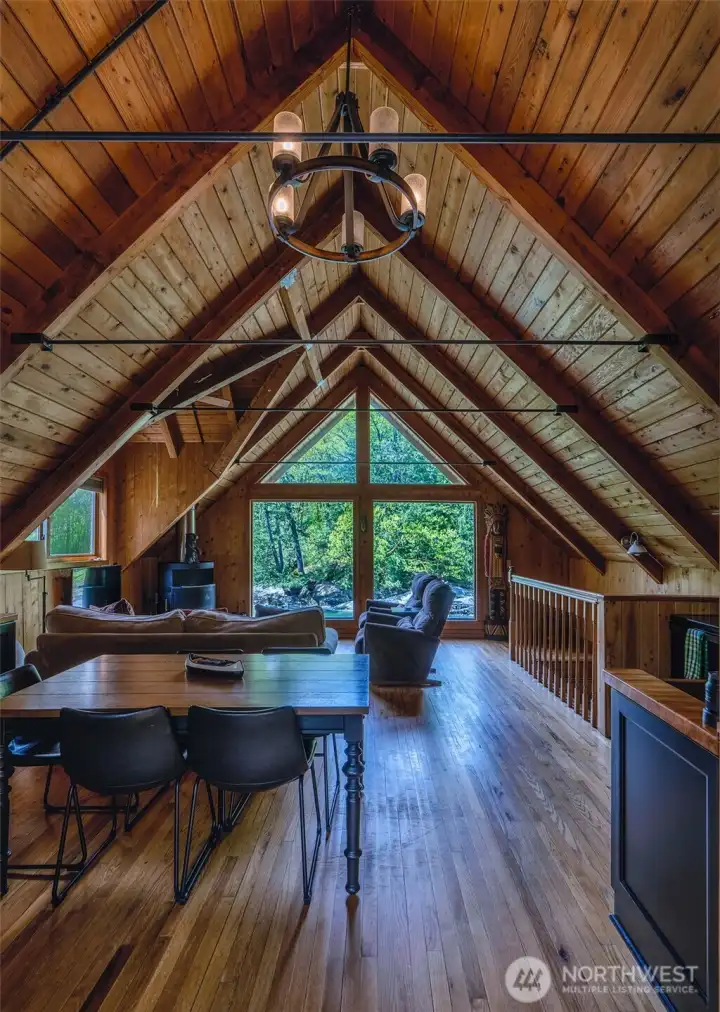 A vaulted great room showcasing open beams, rich wood interior, wood floors, woodstove and an expansive window to soak in the views.