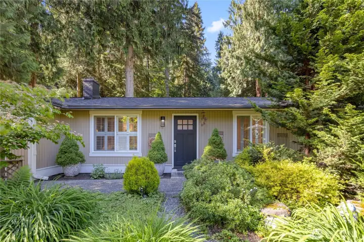 The freshly painted Craftsman-influenced panel door adds a crisp, polished note, finished with a shiny new handle and top-panel glass that brings natural light into the entry. A pretty new cage-style lantern porch light completes the entrance, framed by mature trees, layered plantings, and the quiet retreat feeling that defines this Lake Marcel home.