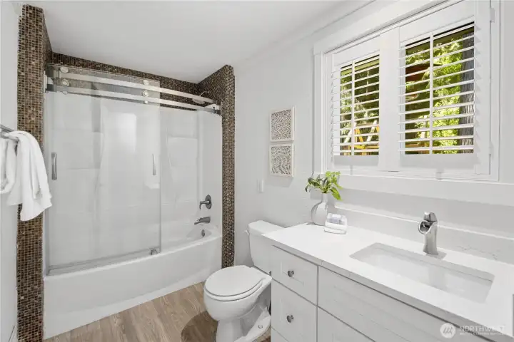 Remodeled in 2024, the guest bath feels fresh and thoughtfully finished, with beautiful copper-toned tile framing the tub/shower and a curved glass sliding door adding a graceful, updated look. The porcelain sink and quartz counter echo the kitchen finishes, while the modern vanity offers generous storage for everyday toiletries. A large window brings in natural light, and just behind the door, a stacking front-load Whirlpool washer and dryer make laundry day easy and convenient.