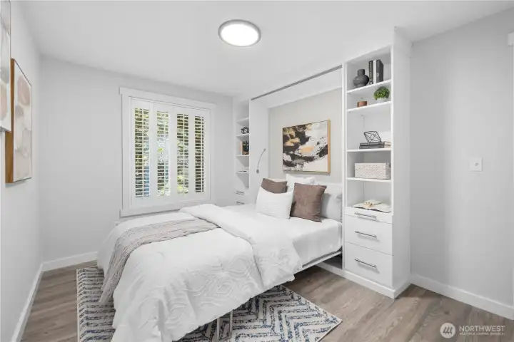 This bedroom offers a west-facing window with classic shutters, the same new luxury laminate hardwood used throughout the home, a large closet, and a Murphy bed framed by built-in shelving for a flexible, space-saving design. The modern flush-mount ceiling light provides an even glow, making the room feel bright, polished, and easy to use as a bedroom, guest room, or multi-purpose space.
