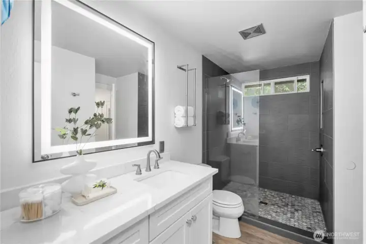 The Jack and Jill primary bath is accessible from both the primary bedroom and the hallway, adding flexibility for everyday use and guests. Quartz counters, white vanity cabinetry, and gray tile tones continue the home’s fresh, cohesive style, with the remodel completed in Dec. 2024. A large vanity mirror features touch-selectable lighting, adding both function and a polished modern look. The walk-in shower is wrapped in floor-to-ceiling tile, with a high window bringing in natural light above, while the glass wall and shower door keep the space feeling open, bright, and beautifully finished.