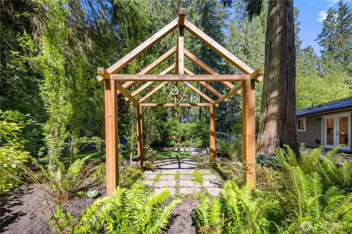 A gorgeous woodland-inspired pergola with a stone-paved floor creates a special place to enjoy the private garden, framed by ferns, flowering plants, and the natural beauty of the yard. It feels like its own little destination within the landscape, perfect for quiet moments, conversation, or simply taking in the peaceful setting.
