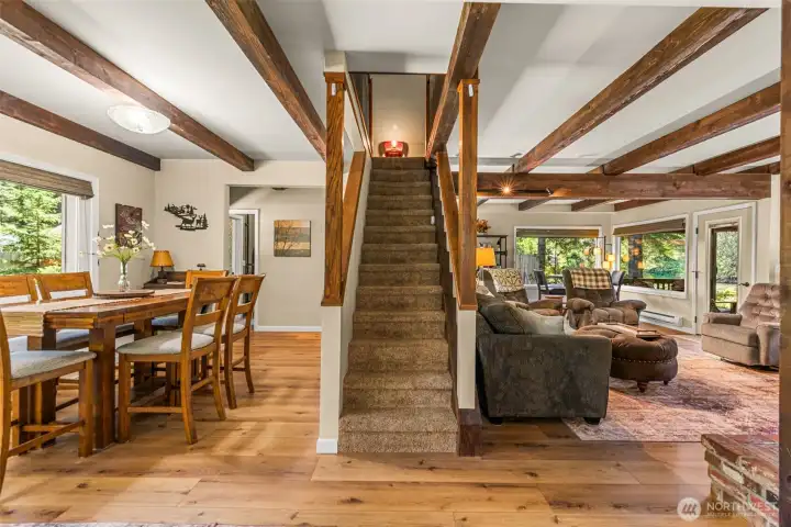 Open-concept great room featuring exposed timber beams, wood flooring, a central staircase, and seamless flow between the dining and living areas Open-concept great room featuring exposed timber beams, wood flooring, a central staircase, and seamless flow between the dining and living areas