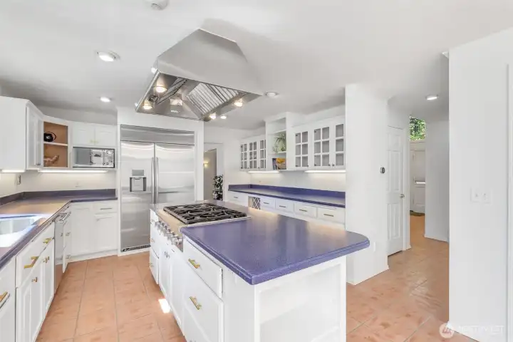 The kitchen extends with abundant counter space, custom cabinetry, and a professional-grade hood above the central island cooktop. Stainless steel appliances and glass-front cabinets add both function and style, while the layout ensures seamless flow for meal preparation and entertaining.