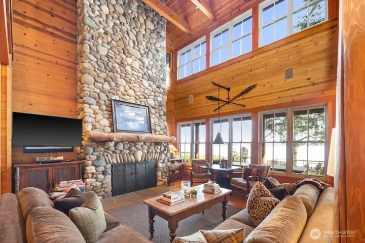 MAGNIFICENT RIVER ROCK FIREPLACE TOWERS ABOVE THE GREAT ROOM FRAMED BY A WALL OF WINDOWS