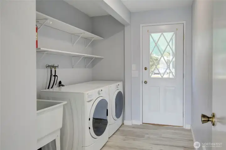 Laundry area offers full-size front-load washer and dryer, a utility sink, built-in shelving for extra storage — all finished with light flooring and fresh gray tones.