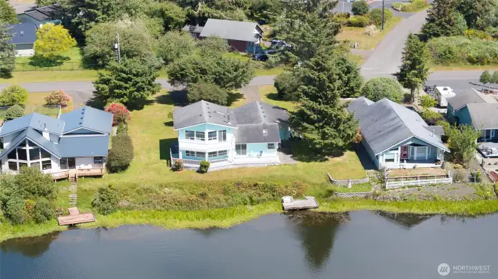 This bird's-eye view captures the property's exceptional canal-front setting — a charming two-story home with a wraparound deck, lush green yard, and private dock right on the water's edge, perfectly positioned to enjoy the tranquil canal and the best of coastal Washington living.