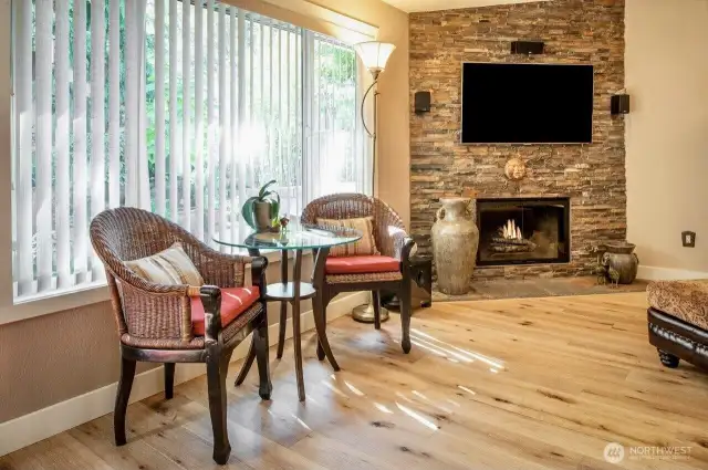 Highlighted by a stunning floor-to-ceiling stacked stone fireplace and mounted TV, perfect for cozy evenings at home. Natural light pours through the oversized window with vertical blinds, creating a bright and airy atmosphere. The elegant bistro nook offers a charming spot for morning coffee or intimate conversation, framed by lush outdoor greenery.