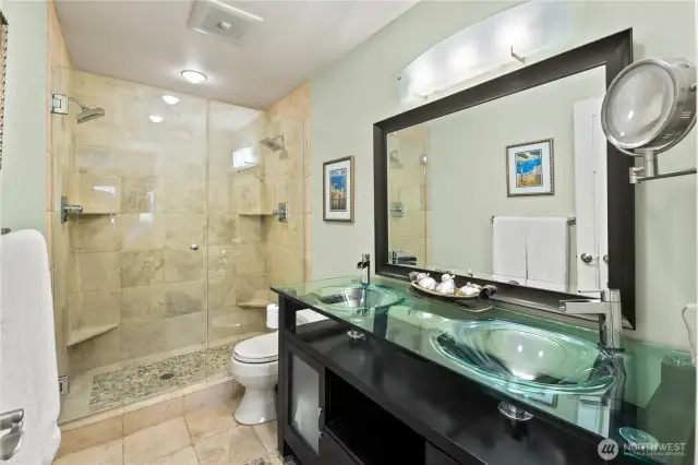 beautifully upgraded primary bathroom offers a spa-like experience with high-end finishes and thoughtful design. The spacious walk-in shower features floor-to-ceiling tile, a built-in bench, and frameless glass doors for a modern, open feel. The double vanity with vessel sinks, glass countertops, and rich espresso cabinetry provides both style and storage, complemented by sleek contemporary lighting.