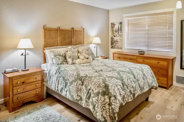 Spacious primary bedroom designed for comfort and relaxation. Natural light filters in through a large window with wood blinds, accentuating the room’s neutral tones and wide-plank hardwood flooring.