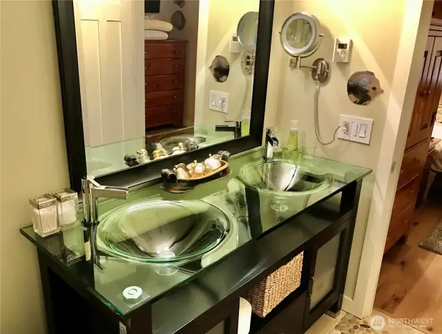 Featuring dual glass vessel sinks, sleek chrome faucets, and a striking glass countertop