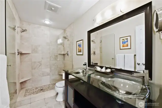 beautifully upgraded primary bathroom offers a spa-like experience with high-end finishes and thoughtful design. The spacious walk-in shower features floor-to-ceiling tile, a built-in bench, and frameless glass doors for a modern, open feel. The double vanity with vessel sinks, glass countertops, and rich espresso cabinetry provides both style and storage, complemented by sleek contemporary lighting.