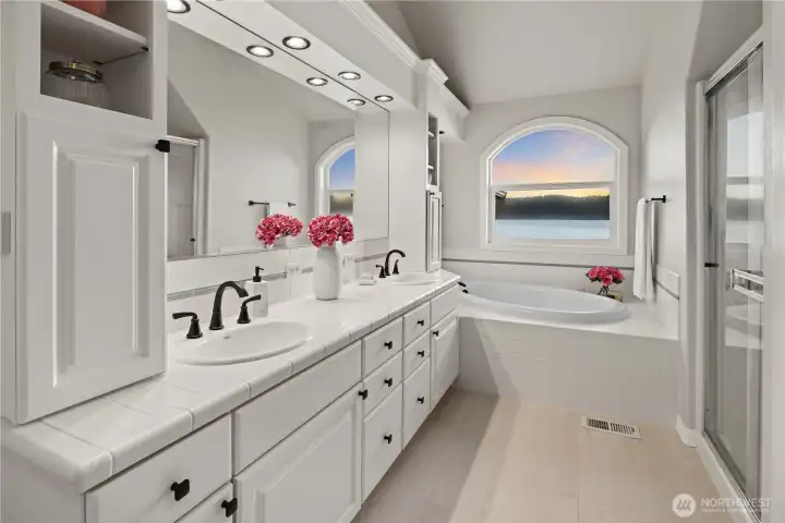 Spacious primary bathroom featuring dual vanities, soaking tub, separate shower, and a beautiful window framing serene waterfront views.