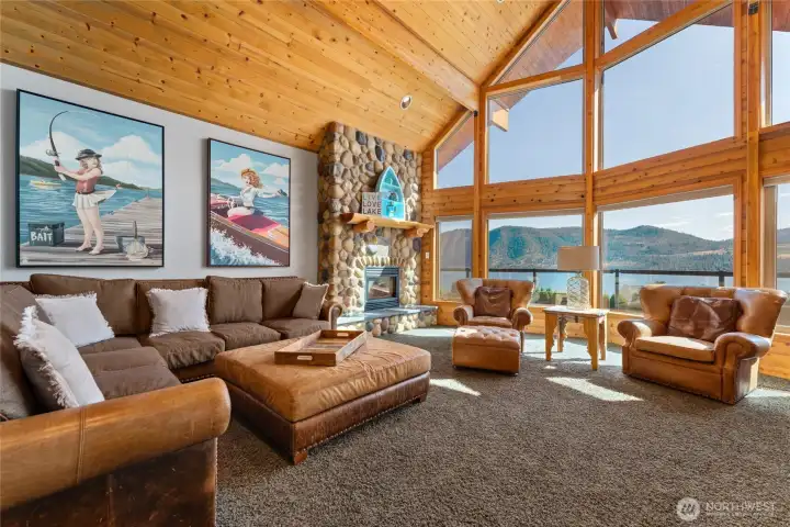 View of of living room w/ oversized windows, river rock fireplace and pine ceilings.