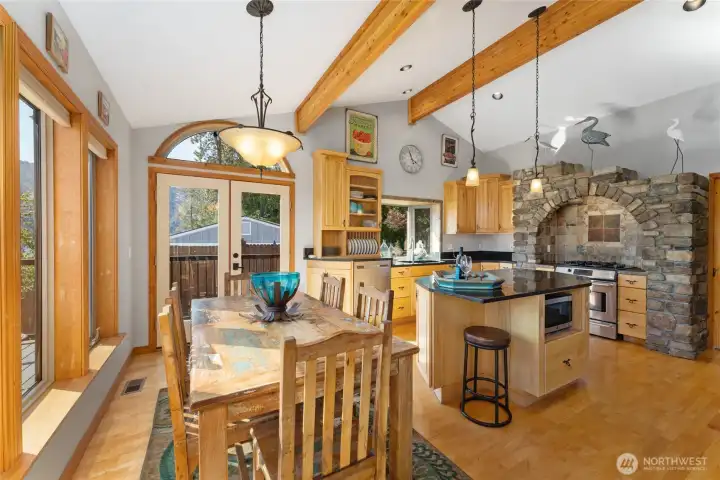 View of dining room and kitchen w/ hardwood floors, stainless steel appliances, quartz countertops, wood beams,  stone surround stove and oven, large windows with views of Lake Chelan and French doors leading to the outdoor living area w/ pool, hot tub, dining and lounge area.