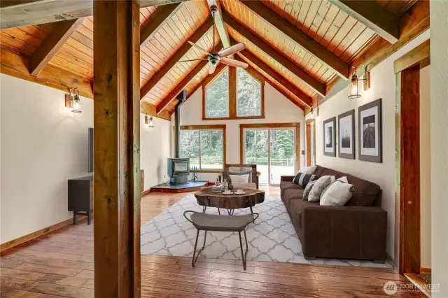 Spacious living area with high, vaulted ceilings made of warm-toned wood planks and exposed wooden beams. A ceiling fan hangs from the peak. The room features a wood stove and large windows and glass doors that look out onto a natural, wooded setting. Virtually staged