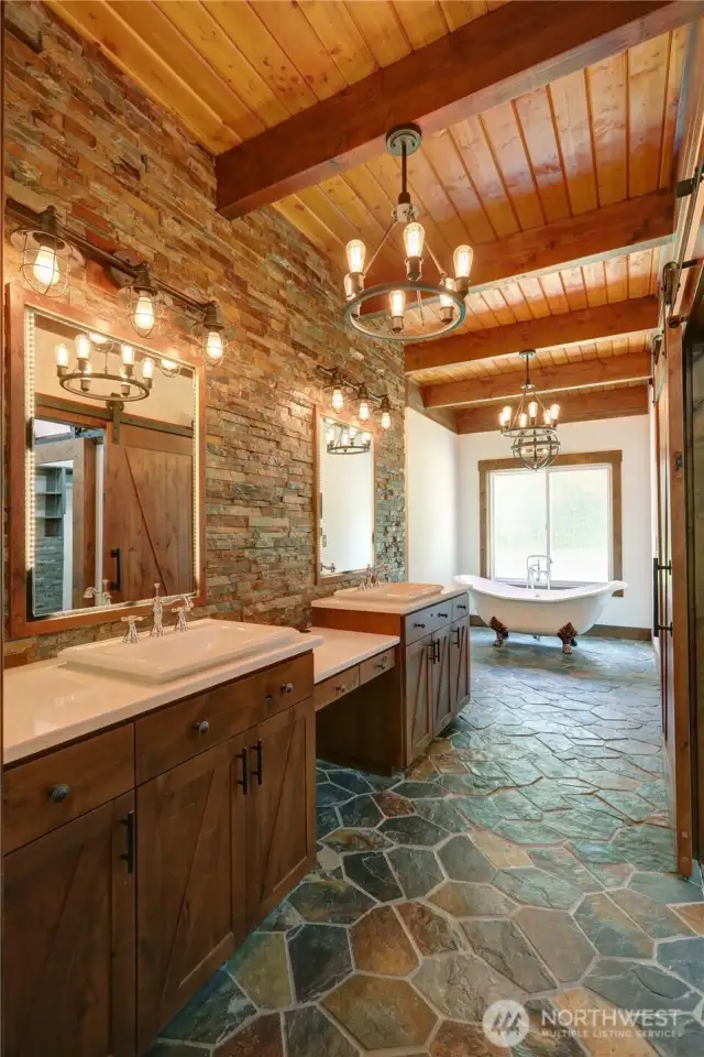 Luxurious primary bathroom with stacked stone veneer, wood plank ceiling with exposed beams, industrial-style light fixtures with exposed bulbs. A freestanding clawfoot bathtub, chandelier, barn style doors, double Sinks, Slate Flooring,