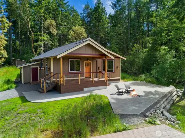 Charming island retreat surrounded by evergreens, featuring a welcoming front porch, spacious patio with firepit, and modern finishes for easy indoor-outdoor living.