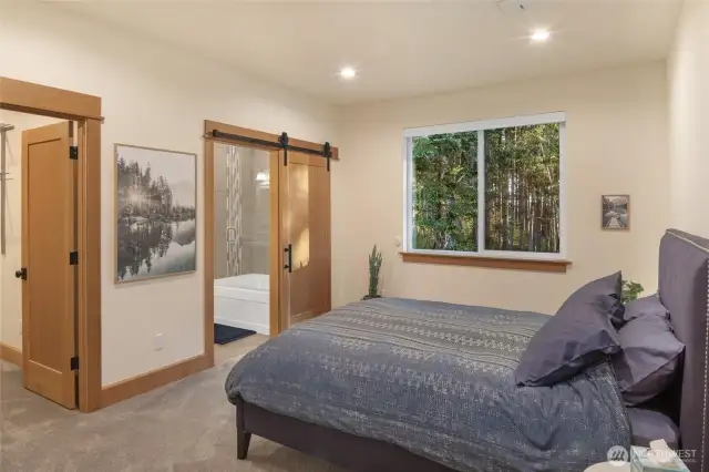 Inviting primary bedroom with serene forest views, cozy carpeting, and ensuite bath with sliding barn door.