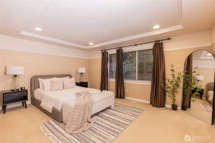 A spacious and serene primary suite featuring tray ceilings, soft ambient lighting, and generous proportions. Large windows invite natural light while warm tones and curated staging create a restful, sophisticated retreat.