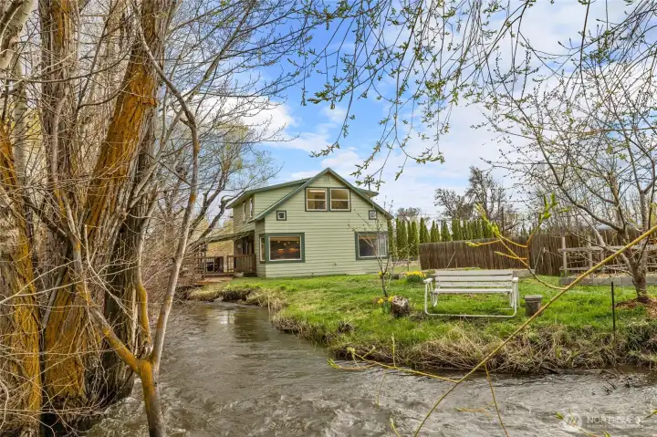 Another view of the home and creek area featuring a cozy seating spot, raised flower beds, and a handy outbuilding for extra storage.