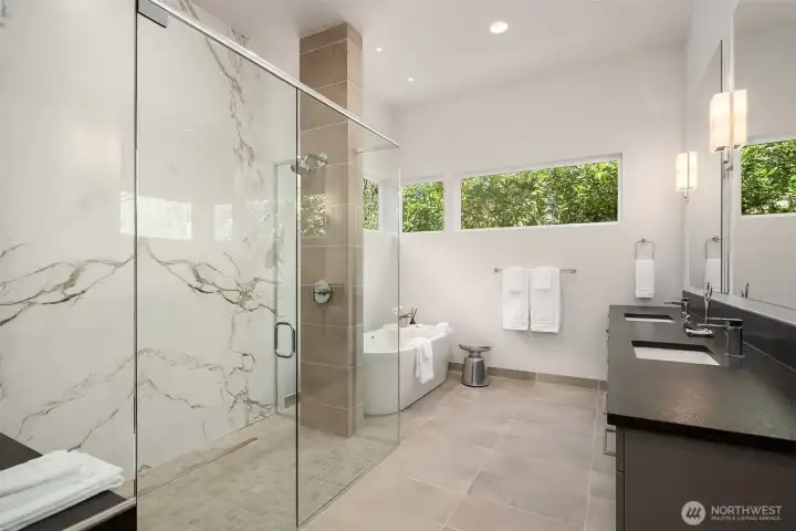 Spa-like primary bath with slab surfaces, walk-in shower, and a freestanding soaking tub framed by greenery.
