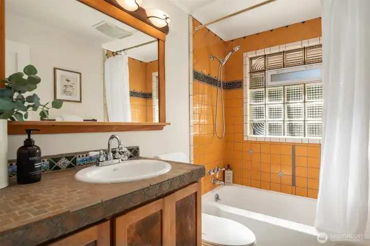 The thoughtfully updated bath blends warmth and craftsmanship with heated travertine floors, a custom cherry vanity with copper details, artisan tilework, and a striking glass block window.