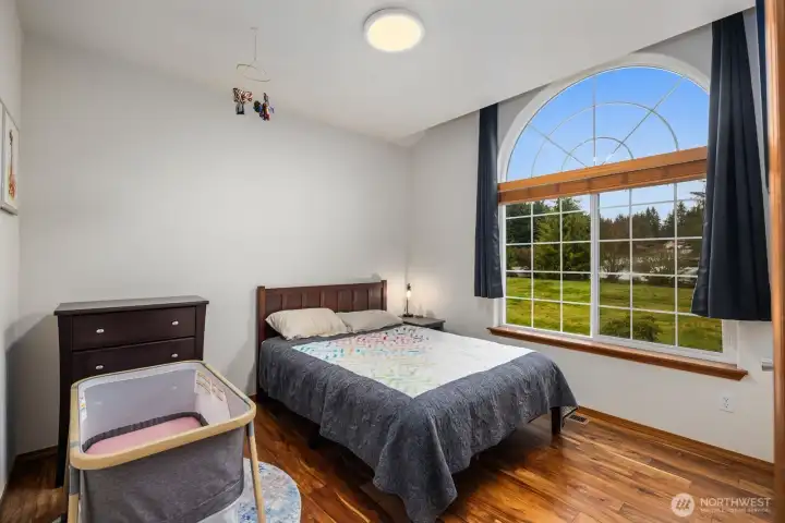 Bedroom on the main level featuring a large arched window that fills the space with natural light and provides views of the surrounding yard and trees.