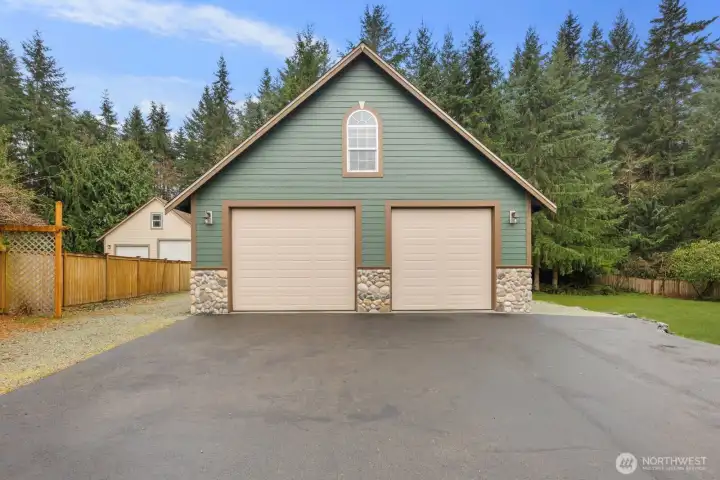 Detached shop building with two large garage bays and additional driveway parking, positioned behind the home and surrounded by mature evergreen trees. Ideal space for vehicles, storage, or workshop use.
