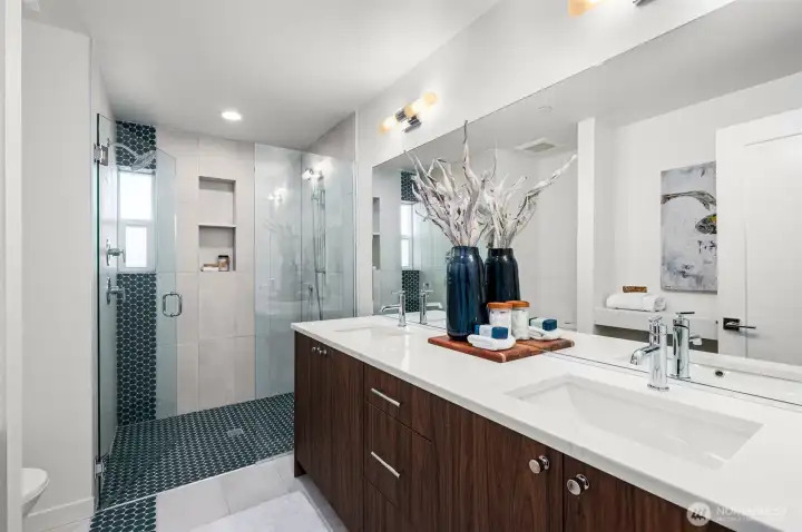 Photos of model home 9213 with similar finishes & floor plan. The primary bath gives a spa like plcae: dual sinks, a walk-in shower, warm tones, and heated floors for extra comfort.